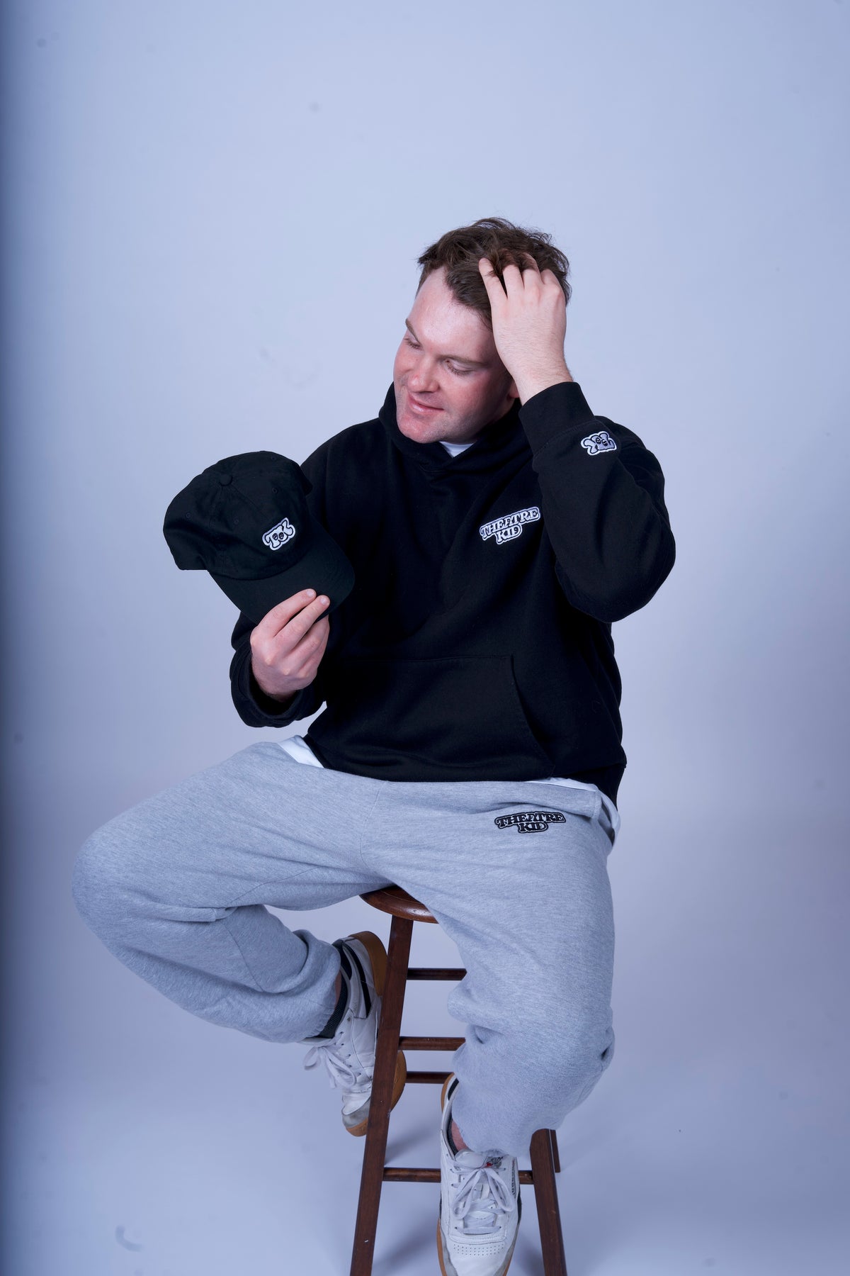 A man sitting on a wooden stool wearing a black Theatre Kid hoodie and light gray Theatre Kid sweatpants, holding a black Theatre Kid baseball cap in his hand against a light gray studio background.